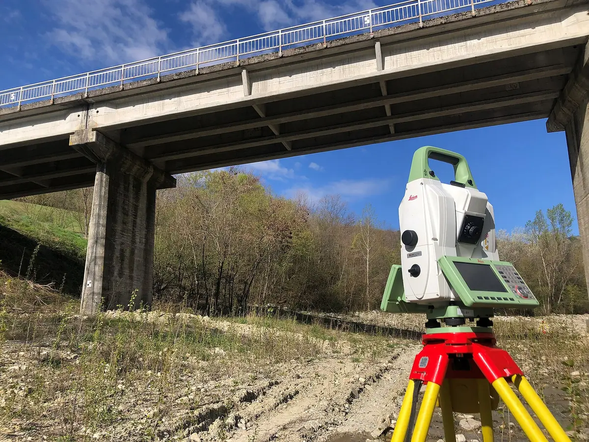 Een totaalstation op een rood-geel statief onder een betonnen brug